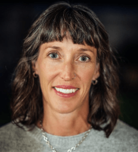 Headshot of Dr. Jennifer Watt wearing light gray sweater with silver necklace. Dr. Watt has bangs and shoulder-length wavy brown hair.