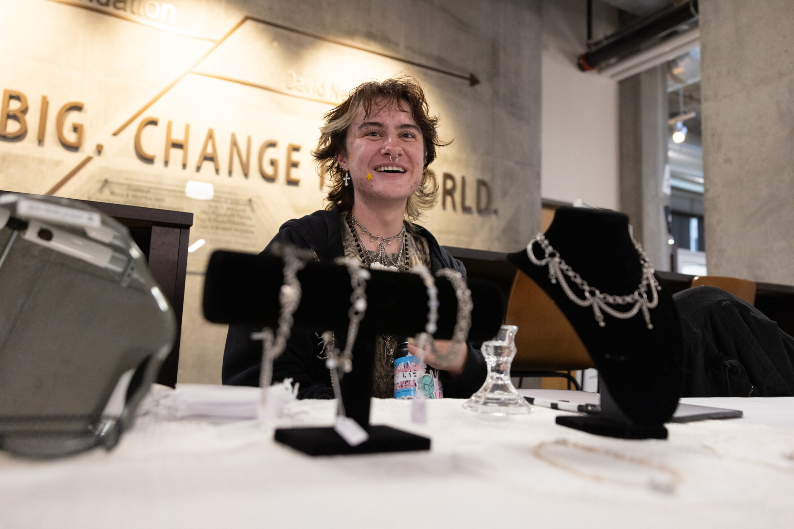 A smiling brunette person is seated at a table behind several velvet jewelry displays.