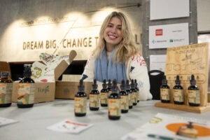 A smiling blond person is seated at a table behind a row of small bottles with dropper tops.